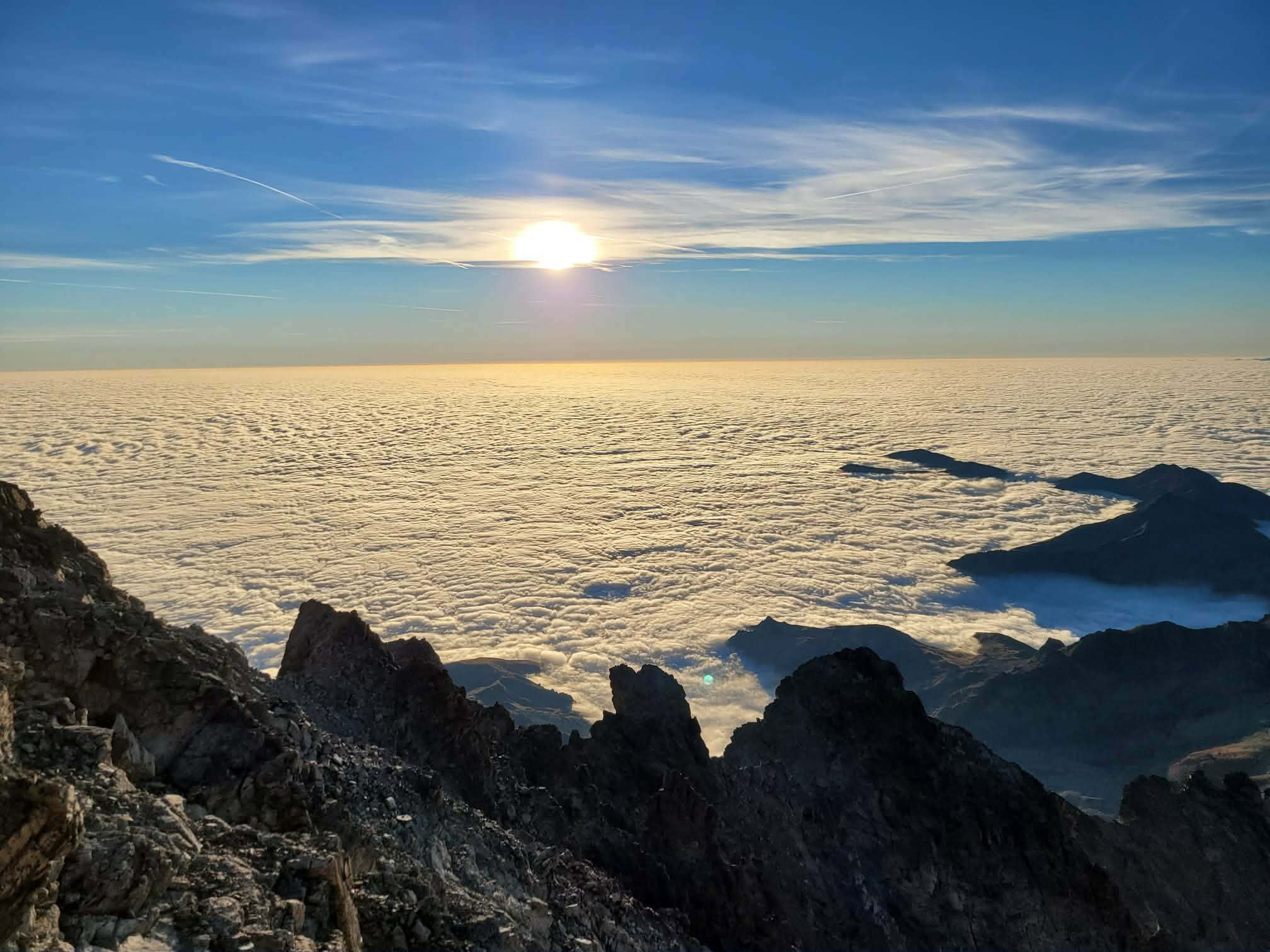 Vue depuis un sommet rocheux au-dessus d’une mer de nuages au lever du soleil, avec l’horizon doré et un ciel bleu dégagé.